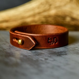 Op de afbeelding: Een bruine leren manchetarmband met een messing knoopsluiting. De armband heeft de tekst "6.0" in reliëf. Het leer heeft een rijke, warme bruine kleur. De manchet is een eenvoudig, maar stijlvol accessoire.