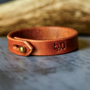 May include: A brown leather bracelet with a brass clasp and the embossed number "60". The bracelet is made of a single band of leather and is designed to be worn around the wrist. The background is a blurred wooden texture.