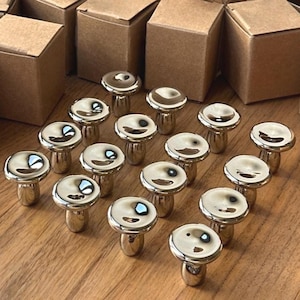 Peut inclure: Plusieurs boutons de meuble argentés en forme de champignon. Les boutons sont disposés sur une surface en bois, avec plusieurs petites boîtes en carton marron en arrière-plan. Les boutons ont une surface lisse et réfléchissante.