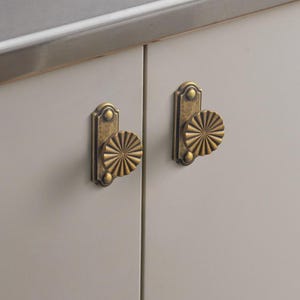 May include: Two antique brass cabinet knobs with a sunburst design. The knobs are mounted on a light grey cabinet door. The knobs have a rectangular backplate with a decorative top and bottom.