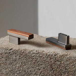 Könnte beinhalten: Zwei moderne Schrankgriffe mit minimalistischem Design. Einer hat eine rechteckige Holzplatte mit einem silbernen Metallsockel. Der andere hat einen rechteckigen Metallkörper mit einem Holzakzent. Beide stehen auf einer strukturierten Steinoberfläche.