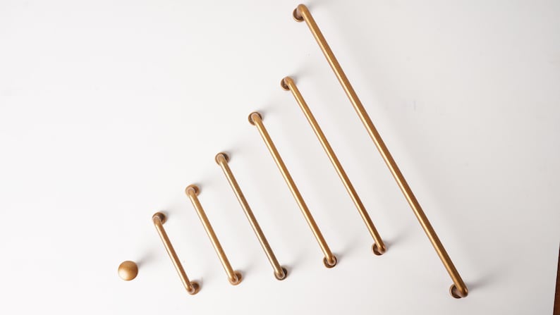 May include: A collection of seven gold-toned cabinet hardware pieces arranged in increasing size order. The set includes one round knob and six bar-style drawer pulls of varying lengths, all featuring a smooth, matte metallic finish.