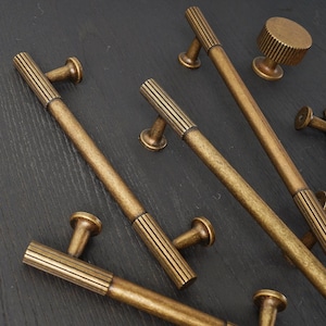 May include: An assortment of antique brass cabinet hardware, including handles and a knob. The handles are long and cylindrical with grooved detailing in the center. The knob is round with a ridged surface. The hardware is arranged on a dark surface.