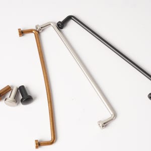 May include: Three long, thin, L-shaped metal cabinet pulls in brass, silver, and black finishes, accompanied by three matching small, cylindrical cabinet knobs. Each pull features a straight bar with a perpendicular mounting post at each end.