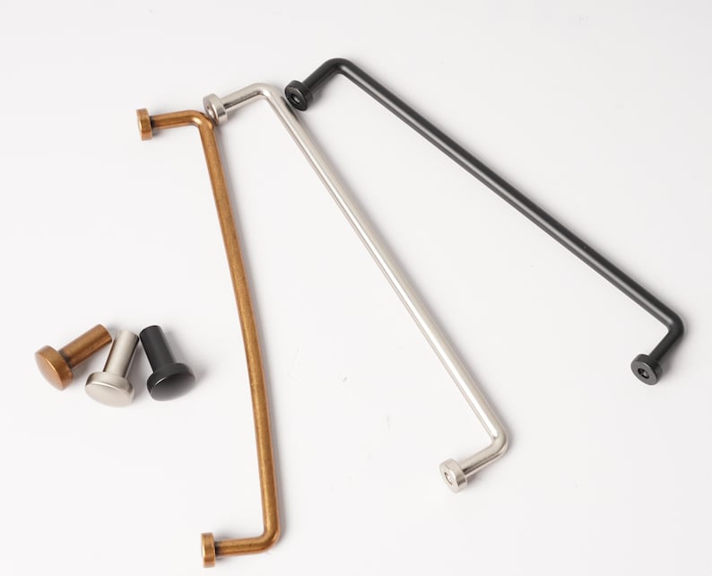 May include: Three long, thin, L-shaped metal cabinet pulls in brass, silver, and black finishes, accompanied by three matching small, cylindrical cabinet knobs. Each pull features a straight bar with a perpendicular mounting post at each end.