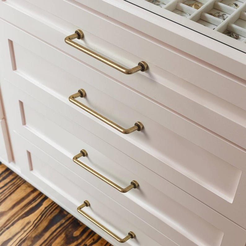 May include: A light pink dresser featuring four horizontal drawers, each equipped with a slim, antique brass-toned metal pull handle. The top drawer is partially open, revealing a white jewelry organizer tray with multiple small compartments.