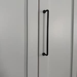May include: A long, matte black metal cabinet pull handle with a cylindrical bar design and two circular mounting points, attached vertically to a white cabinet door with a recessed panel frame and a smooth, clean finish.