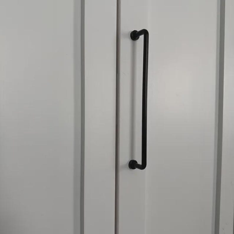May include: A long, matte black metal cabinet pull handle with a cylindrical bar design and two circular mounting points, attached vertically to a white cabinet door with a recessed panel frame and a smooth, clean finish.