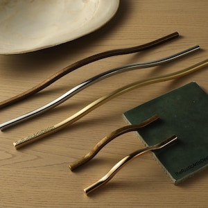 Op de afbeelding: Een verzameling golvende kastgrepen in verschillende afwerkingen: donker hout, zilver, goud en messing. De grepen zijn gerangschikt op een licht houten oppervlak, samen met een groene vierkant en een zeeschelp. The RusticArtisticDesign is gedrukt.