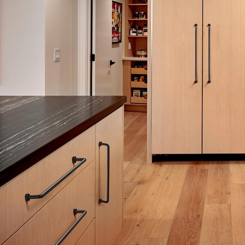 May include: A set of matte black metal cabinet pulls and handles installed on light-colored wood cabinetry. The hardware features a cylindrical bar design with rounded ends, providing a contrast against the light wood grain of the kitchen drawers and tall pantry doors.