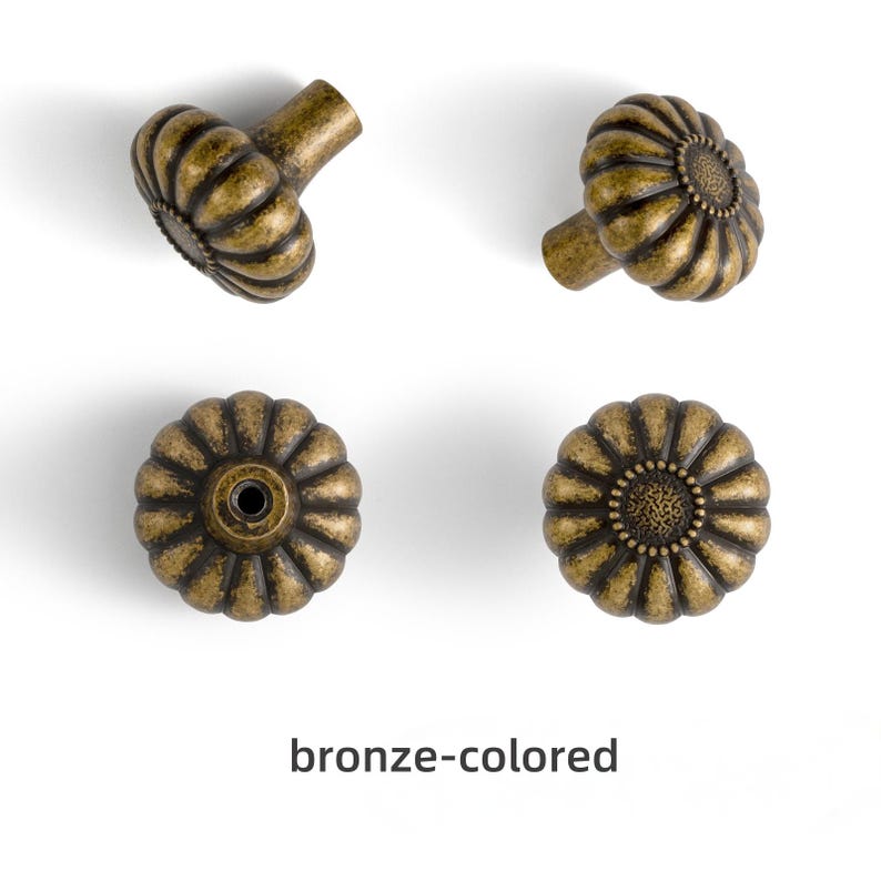 May include: Four bronze-colored cabinet knobs with a round, fluted, flower-like shape. Each knob features a textured center and a metallic finish with darker highlights in the grooves. The text "bronze-colored" is printed below the knobs.