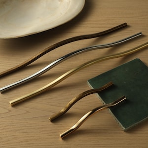 May include: An assortment of wavy cabinet pulls in various finishes: dark brown, silver, and gold. Two smaller pulls are also visible. A green square is next to the pulls. The pulls are on a light wood surface.