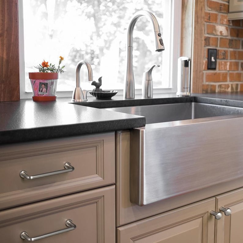 May include: A stainless steel farmhouse sink with a high-arc pull-down faucet and a secondary smaller faucet. The kitchen features light taupe cabinets with silver bar pulls, a black countertop, and a small potted plant with orange flowers.