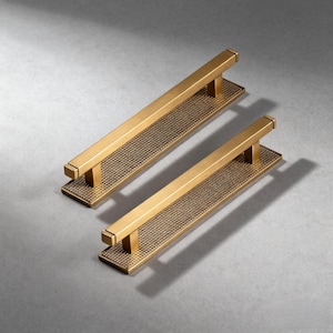 Peut inclure: Deux poignées de meuble en laiton. Chaque poignée a une base rectangulaire avec un motif texturé en forme de grille. Une barre rectangulaire s'étend vers le haut depuis la base, formant la poignée. Les poignées sont de tailles différentes, la plus grande mesurant environ 30 cm de long.