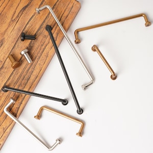 May include: A collection of metal cabinet hardware including long bar pulls and small cylindrical knobs in matte black, brushed silver, and antique brass finishes, arranged in a scattered layout with some pieces resting on a wooden board.