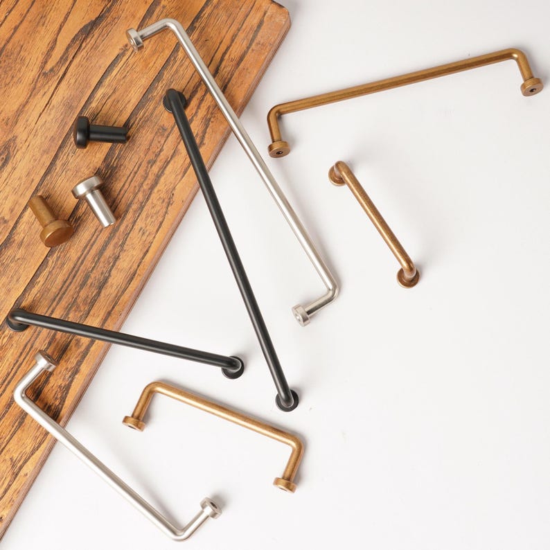May include: A collection of metal cabinet hardware including long bar pulls and small cylindrical knobs in matte black, brushed silver, and antique brass finishes, arranged in a scattered layout with some pieces resting on a wooden board.
