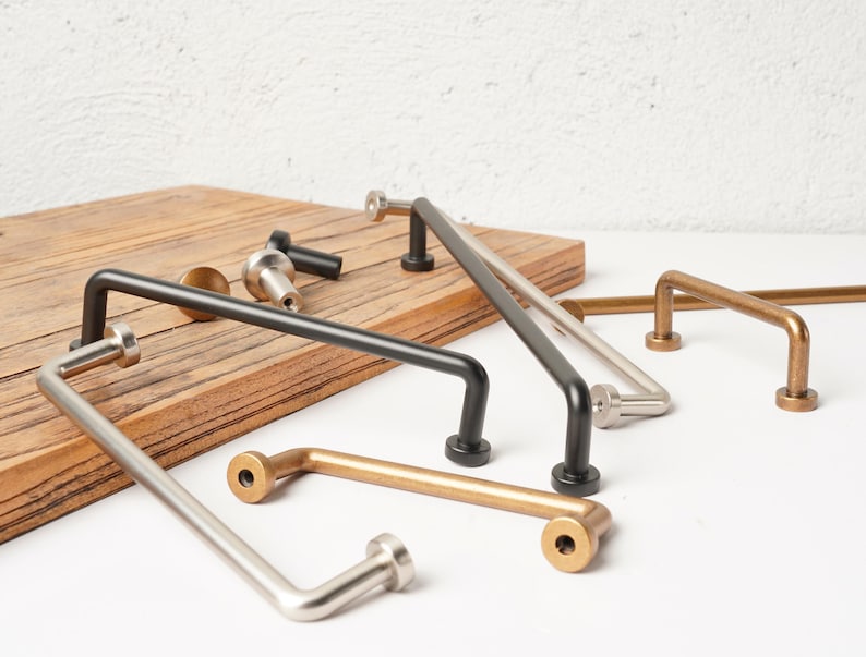 May include: A collection of metal cabinet pulls and knobs in various finishes including matte black, brushed nickel, and antique brass. These hardware pieces feature a simple U-shaped design with cylindrical mounting bases for attachment.