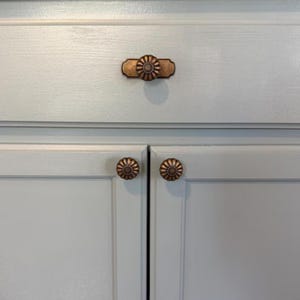 May include: A set of three antique copper-toned metal cabinet knobs installed on light blue painted wooden cabinetry. One knob features a rectangular backplate with a circular fluted design, while the two lower knobs are circular and fluted.