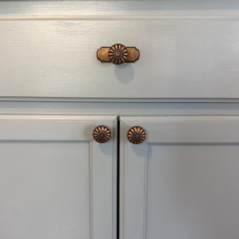 May include: A set of three antique copper-toned metal cabinet knobs installed on light blue painted wooden cabinetry. One knob features a rectangular backplate with a circular fluted design, while the two lower knobs are circular and fluted.