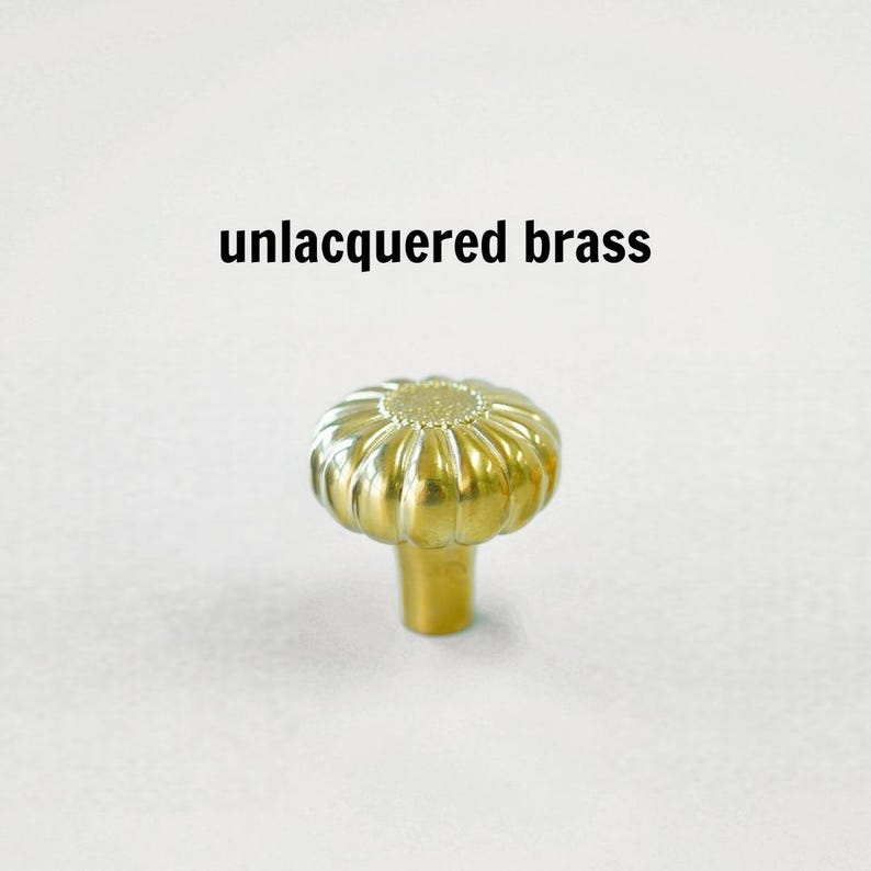 May include: A gold-colored unlacquered brass cabinet knob with a rounded, fluted, pumpkin-like shape and a textured center. The text 'unlacquered brass' is printed above the knob, which features a smooth, metallic finish and a cylindrical base.