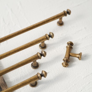 Puede incluir: Primer plano de varios tiradores y manijas de gabinete de latón antiguo. Los tiradores son largos y en forma de barra, mientras que las manijas tienen forma de T. El herraje tiene un aspecto vintage y envejecido, con un color marrón dorado cálido. El diseño presenta detalles decorativos.