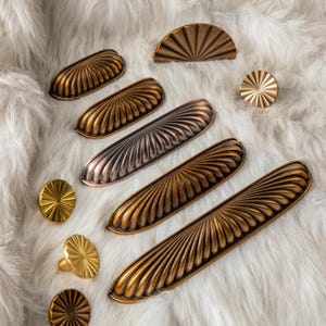 Puede incluir: Una variedad de herrajes decorativos para armarios en varias formas y acabados. La colección incluye tiradores y pomos en tonos bronce, cobre y dorado con un diseño en forma de abanico. Los herrajes están dispuestos sobre una superficie blanca y esponjosa.