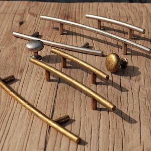 May include: A collection of cabinet hardware, including silver and gold-toned handles and knobs. The handles are curved and straight, with a textured finish. The knobs are round and have a similar aged appearance. The hardware is displayed on a wooden surface.