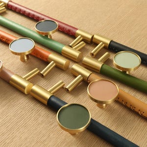 Puede incluir: Una colección de tiradores de armario con herrajes de latón y mangos de cuero o madera pintada. Los tiradores presentan una variedad de colores, incluyendo marrón, verde y negro, y están dispuestos sobre una superficie marrón claro.
