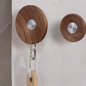 Op de afbeelding: Twee ronde houten wandhaken met een zilverkleurig metalen midden. Aan één haak hangt een houten keukengerei. De haken zijn bevestigd aan een witte muur.