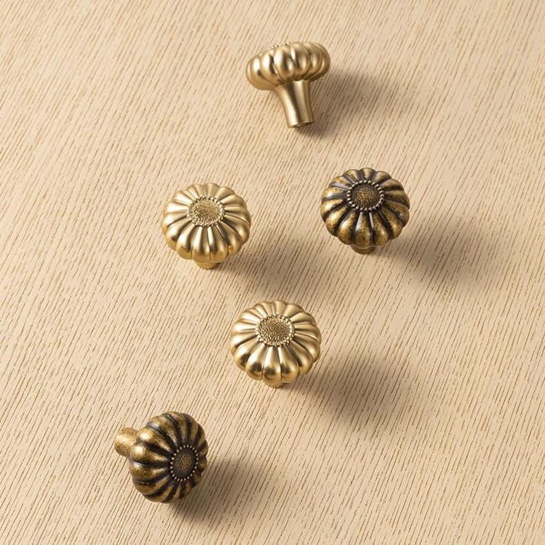 May include: Five round cabinet knobs with a metallic gold finish. Each knob features a fluted, pumpkin-like shape with a textured, circular center detail. The knobs are arranged in a scattered pattern, showing both top-down and side-angle views.