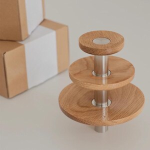 Puede incluir: Un soporte de servicio de tres niveles con plataformas circulares de madera de diferentes tamaños, sostenidas por un poste central de metal plateado. El soporte está hecho de madera clara y metal. Dos cajas de cartón están en el fondo.