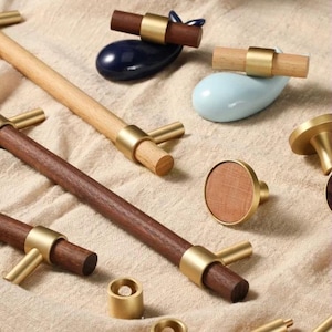 Pode incluir: Uma coleção de ferragens para armários em madeira e latão, incluindo puxadores e puxadores. Os puxadores apresentam cabos cilíndricos de madeira com detalhes em latão. Alguns puxadores têm uma face redonda de madeira. O hardware é exibido em um tecido bege.