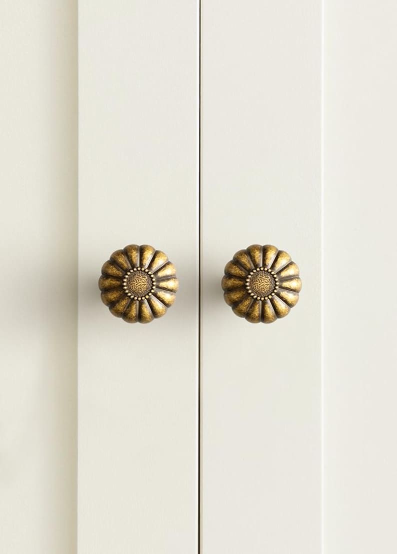 May include: Two round, antique brass-finished cabinet knobs with a flower-like, scalloped petal design. Each knob features a textured, circular center surrounded by radiating, rounded ridges, mounted on a smooth, off-white vertical panel.
