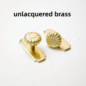 May include: Two unlacquered brass door knobs with a floral, fluted design, each mounted on a rectangular backplate with rounded corners. The metal has a warm, golden tone and a smooth, polished finish. The text unlacquered brass is visible.