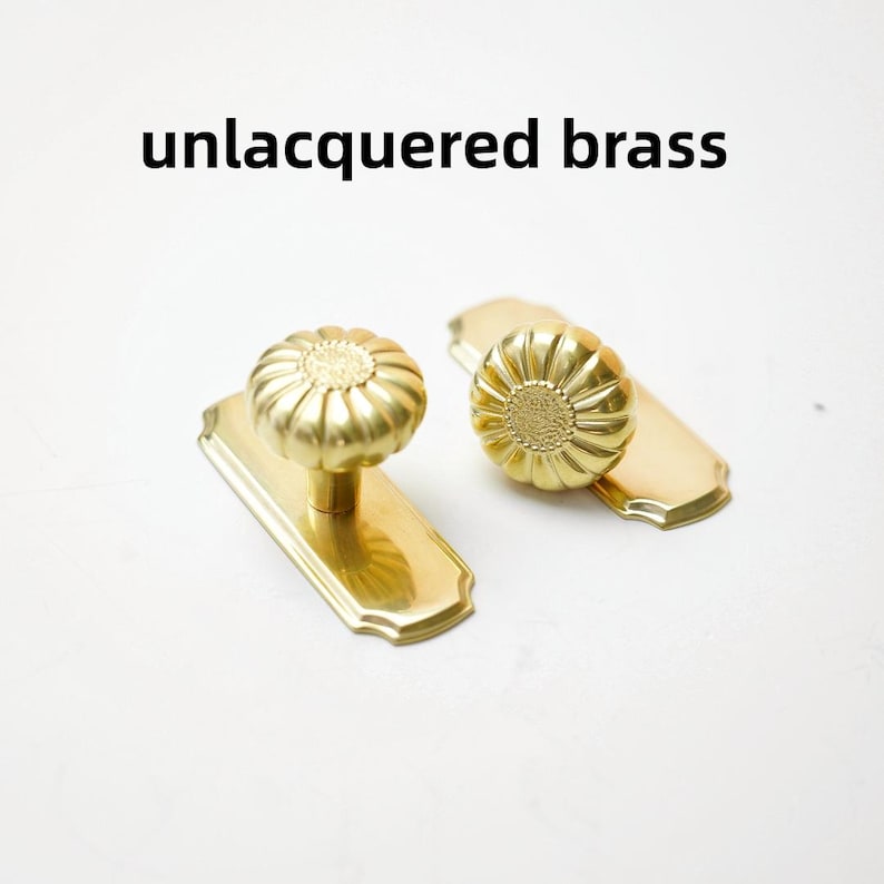 May include: Two unlacquered brass door knobs with a floral, fluted design, each mounted on a rectangular backplate with rounded corners. The metal has a warm, golden tone and a smooth, polished finish. The text unlacquered brass is visible.