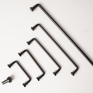 May include: A collection of five matte black metal cabinet pulls of varying lengths arranged in a diagonal line, accompanied by a single matching black cabinet knob positioned to the left of the smallest handle, all featuring a simple L-shaped design.
