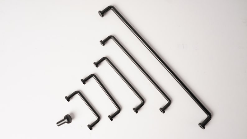 May include: A collection of five matte black metal cabinet pulls of varying lengths arranged in a diagonal line, accompanied by a single matching black cabinet knob positioned to the left of the smallest handle, all featuring a simple L-shaped design.