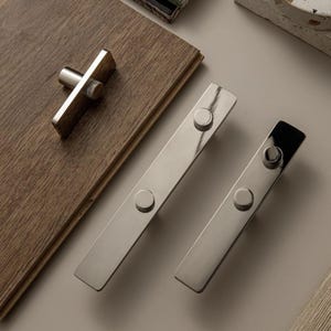May include: A selection of contemporary cabinet hardware in a brushed nickel finish. The set features two long rectangular pulls with circular details and a T-shaped knob. The hardware is presented on a light grey surface and a wooden surface.