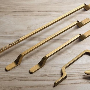 May include: A collection of brass-toned cabinet hardware, including four handles of varying lengths and a small knob. The handles have a distinctive curved design and are arranged on a light wood-grain surface. The text "TheRusticArtsDesign" is visible.