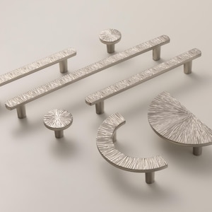 Puede incluir: Una colección de herrajes plateados para armarios, que incluye tiradores de barra, pomos y un tirador curvo. El herraje presenta un diseño texturizado y lineal. Las piezas están dispuestas sobre un fondo claro, mostrando sus diversas formas y tamaños.