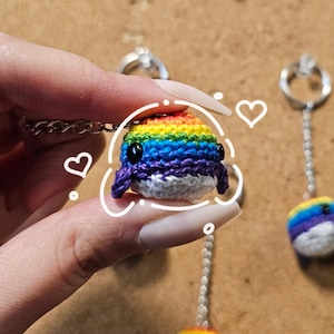 Può includere: Un ciondolo a forma di balena fatto a mano all'uncinetto nei colori dell'arcobaleno, con occhi di perline nere e pinne viola. La balena è attaccata a una catena d'argento. Altri ciondoli a forma di balena arcobaleno sono visibili sullo sfondo.