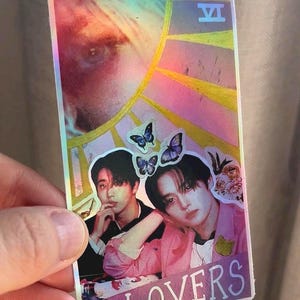 May include: A holographic tarot card featuring two people and the words "THE LOVERS." The card has a VI symbol and butterfly stickers. The top of the card has the text "@the_Doomwitch."