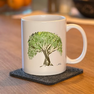 Könnte beinhalten: Weiße Keramiktasse mit einem grünen Baumdesign, auf einem grauen quadratischen Untersetzer. Der Baum hat einen braunen Stamm und grünes Laub. Die Tasse steht auf einem Holztisch, mit verschwommenem Küchenhintergrund.