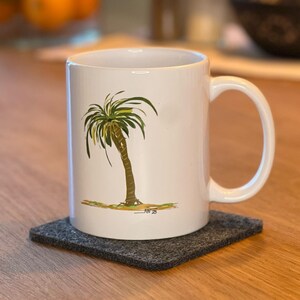 Könnte beinhalten: Weiße Keramik-Tasse mit einer Palmen-Illustration, platziert auf einem dunkelgrauen Filzuntersetzer. Im Hintergrund ist ein Glas mit Orangen und Limetten zu sehen. Die Tasse ist von Standardgröße.