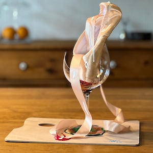 Könnte beinhalten: Ein Paar Ballettschuhe mit hellrosa Band in einem Weinglas. Das Glas steht auf einer Holzpalette mit einem gemalten Design. Der Hintergrund zeigt einen Holztisch und einen Schrank.