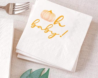 May include: White paper napkins with a pumpkin illustration and the text "oh baby!" in yellow. The napkins are stacked on a light-colored surface, with a silver fork and green leaves nearby. The napkins have a textured pattern.