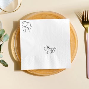 May include: White square napkin with a black line drawing of two balloons and the text "Olivia is 30". The napkin is on a clear plate with a gold fork to the right.