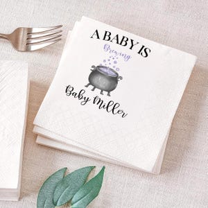 May include: White paper napkins with the text "A Baby Is Brewing" and "Baby Miller" in black script. A gray cauldron with purple bubbles is in the center. A silver fork is in the upper left corner.