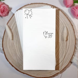 May include: A stack of white paper napkins with a black line drawing of two balloons and the text "Olivia is 30". The napkins are arranged on a wooden slice, with pink roses in the background.