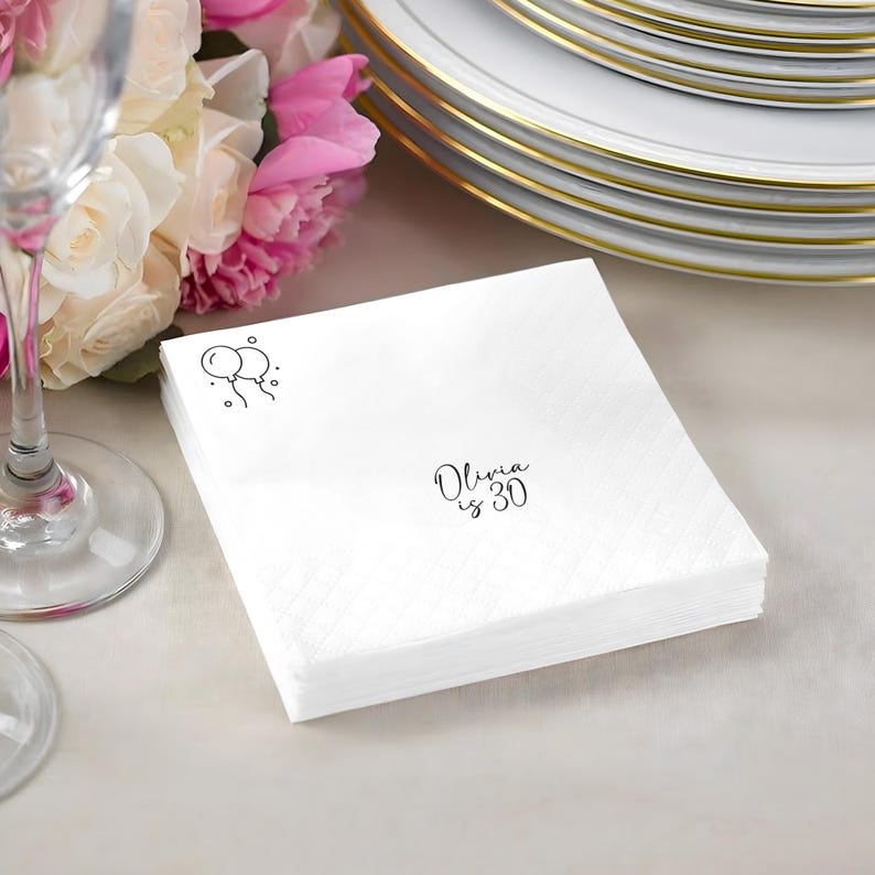 May include: A stack of white paper napkins with a subtle diagonal pattern. The napkins are printed with a small balloon graphic and the text "Olivia is 30" in black script. A stack of white plates with gold rims are in the background.